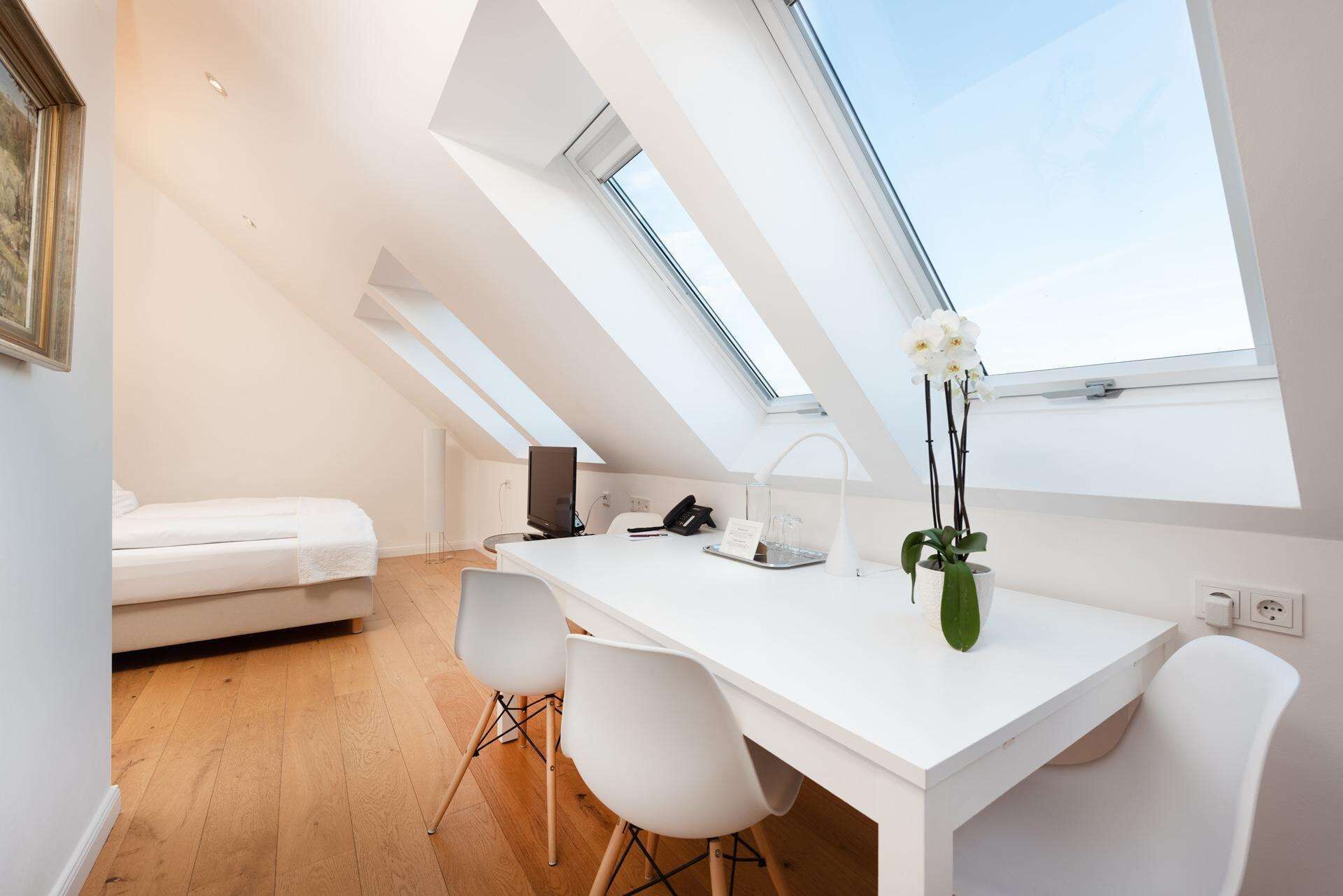 A modern, minimalist room with a sloped ceiling, a white desk, two white chairs, a bed, and a skylight.
