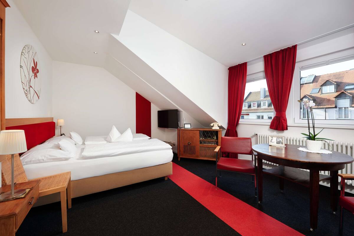 A hotel room with a bed, a desk, a chair, a TV, and a window with red curtains.