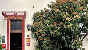 Un hôtel avec une enseigne indiquant «Hôtel Vasteen», un grand arbre aux fleurs jaunes et un chemin en briques menant à l’entrée.