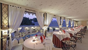 A series of round tables with white tablecloths and black metal chairs, arranged in a covered outdoor area with a view of buildings in the background. - Rohet Garh (Rohat)