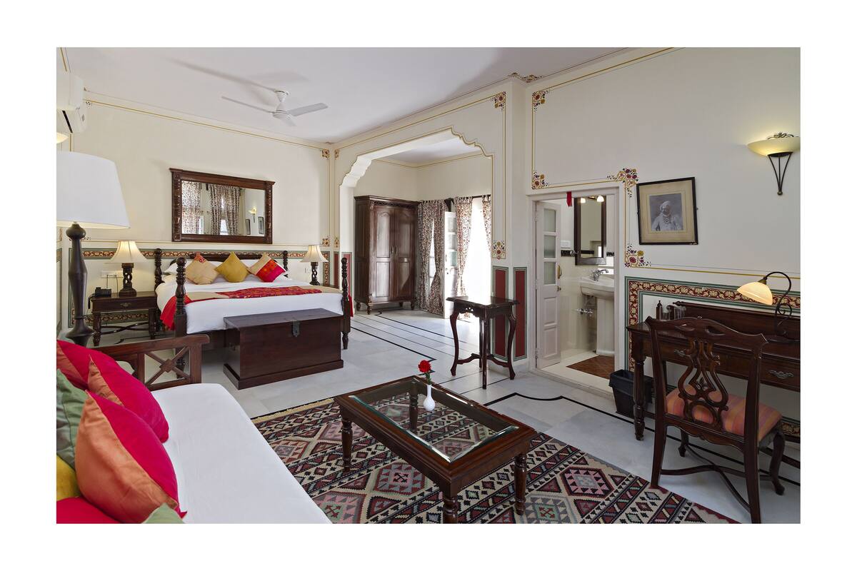 Habitación de hotel con cama, zona de estar con sofá y sillón, mesa de comedor y espejo.