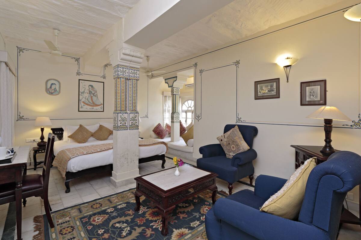 Una habitación tradicional con una cama, dos sillones azules, una mesa de centro y una alfombra estampada.