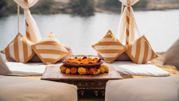 A bed with striped pillows and a bowl of marigold flowers on a wooden table. - Rohet Garh (Rohat)