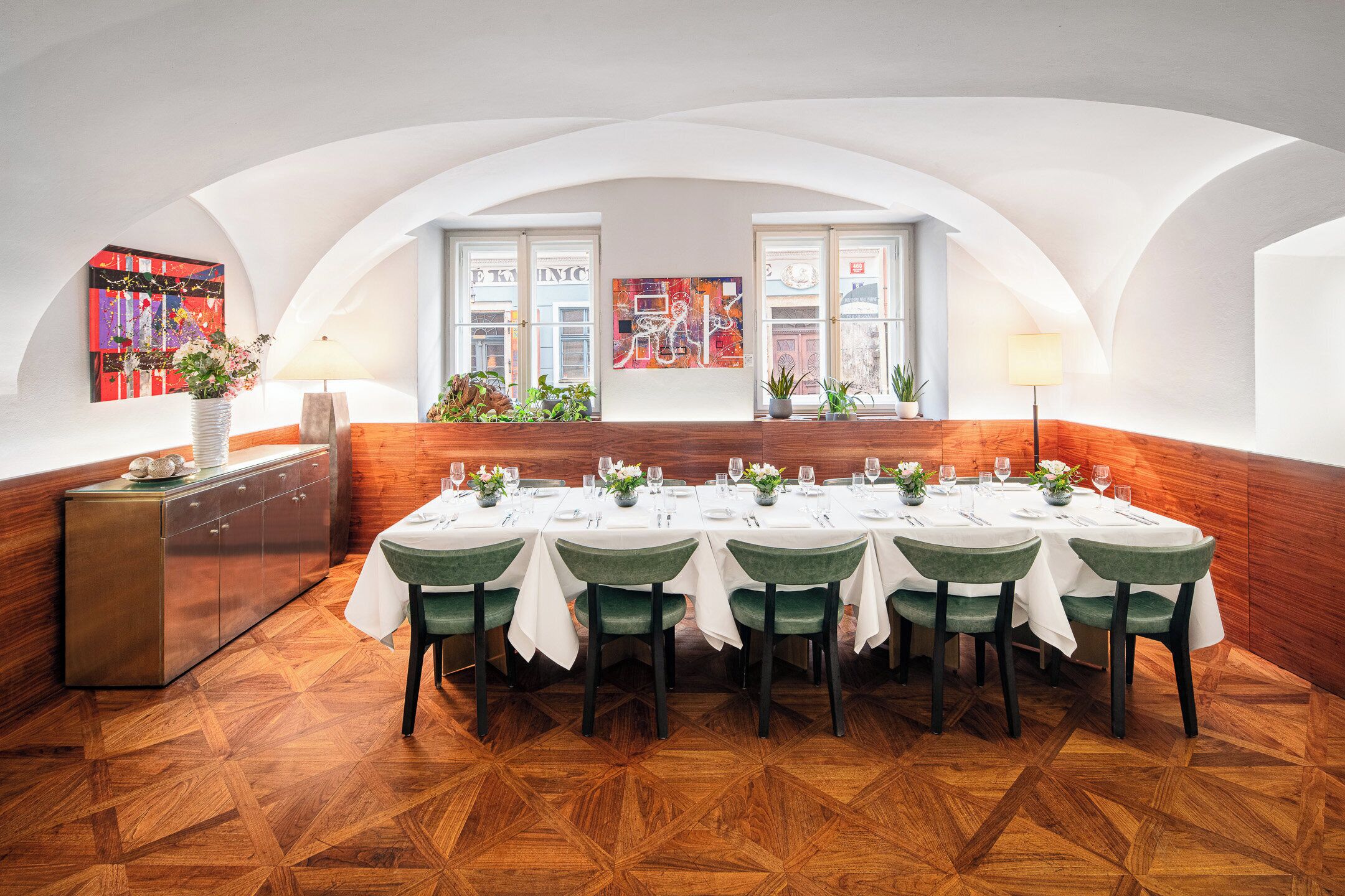 A dining area with a long table set for multiple guests, wooden paneling on the walls, and a view through the windows.