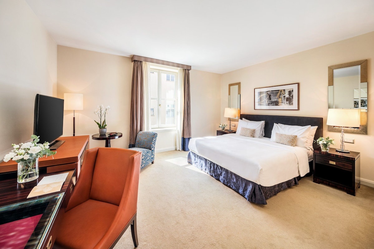 A hotel room with a bed, a desk with a computer, a chair, a nightstand, and a window with curtains.