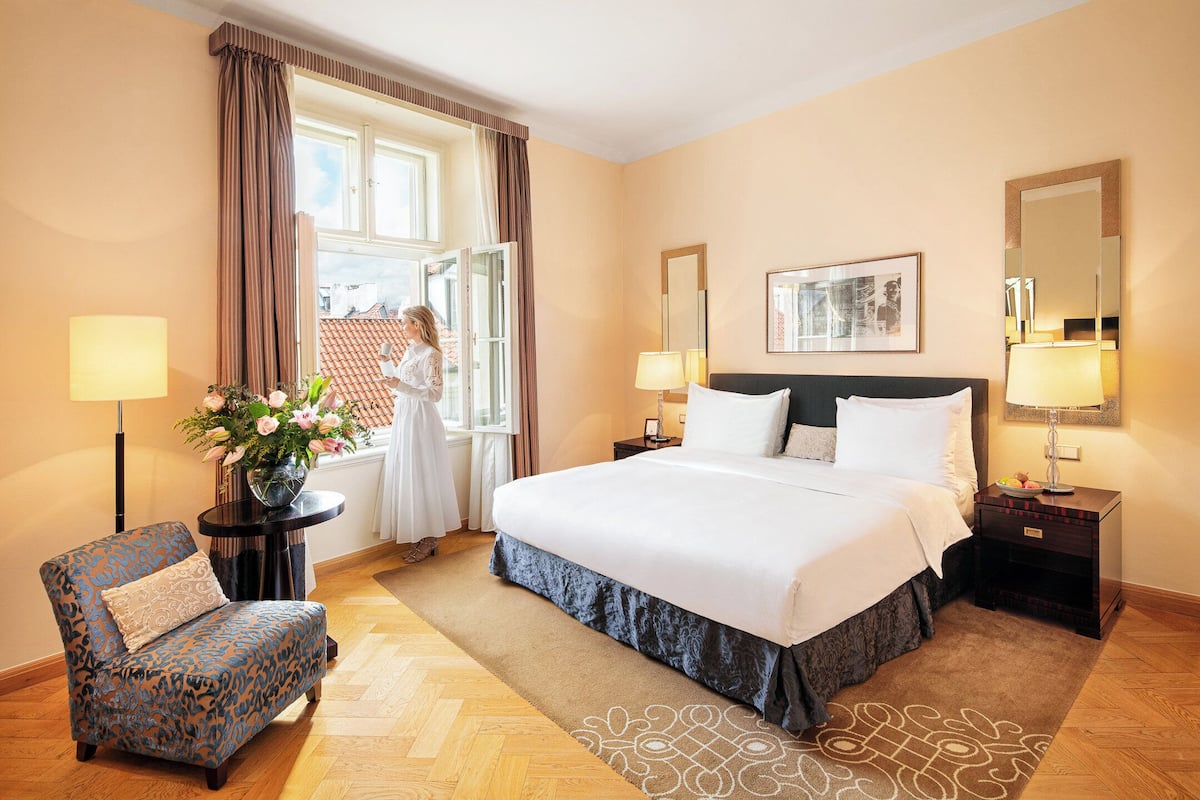 A hotel room with a large bed, a chair, a small table with a lamp, a mirror, and a window with curtains.