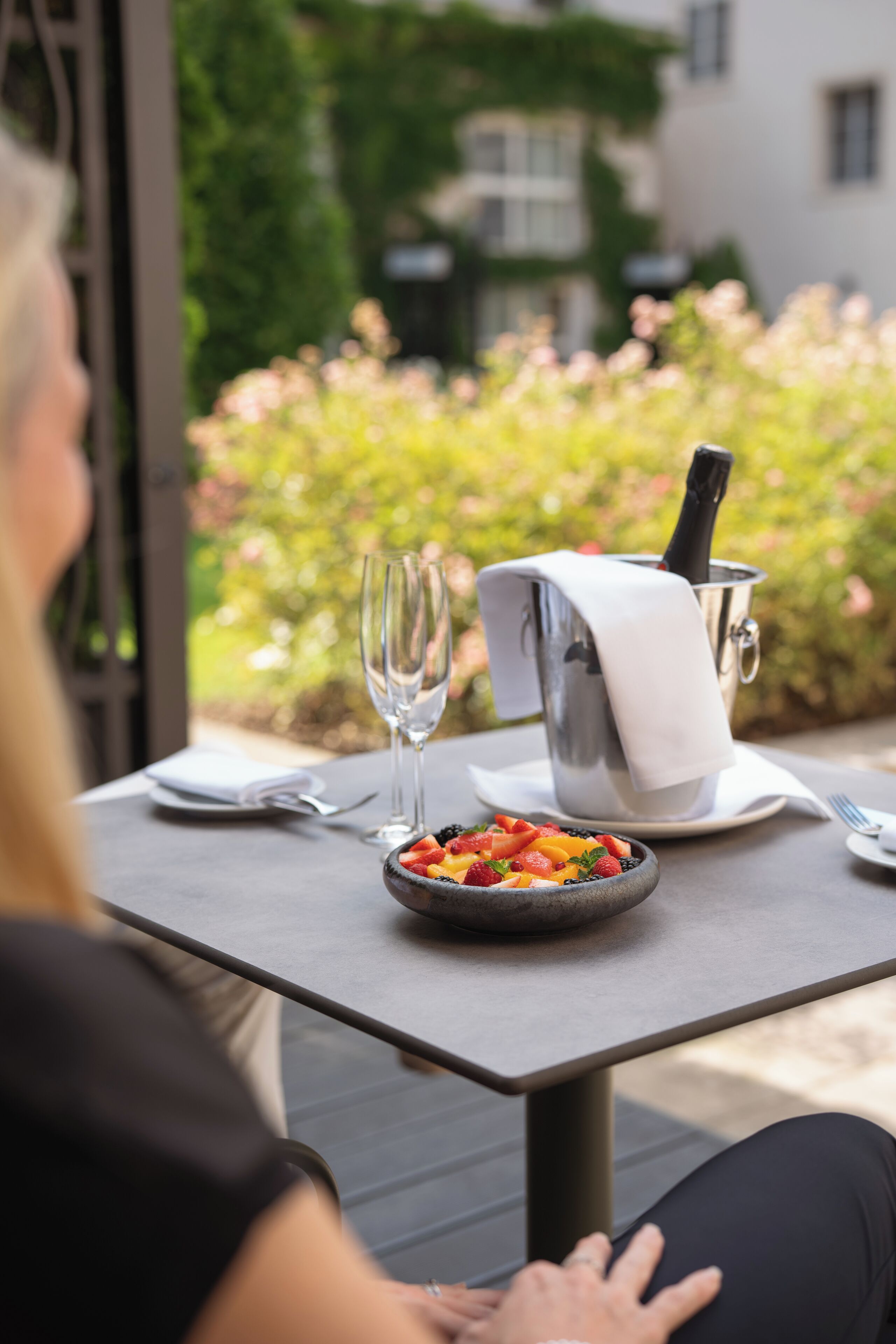 Et bord med en skål frugt, champagneflaske og glas, placeret udendørs med grøn vegetation i baggrunden.