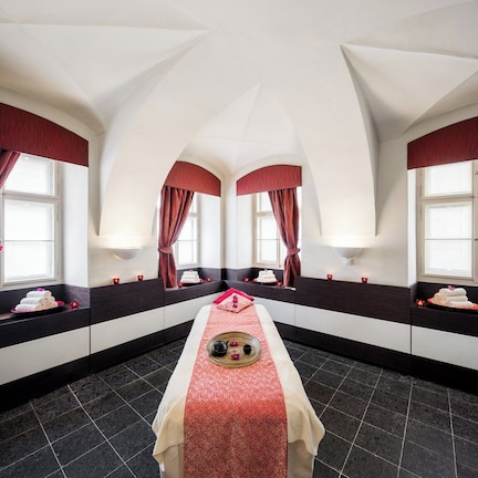 A room with a central bed, two windows with red curtains, and a dark tiled floor.