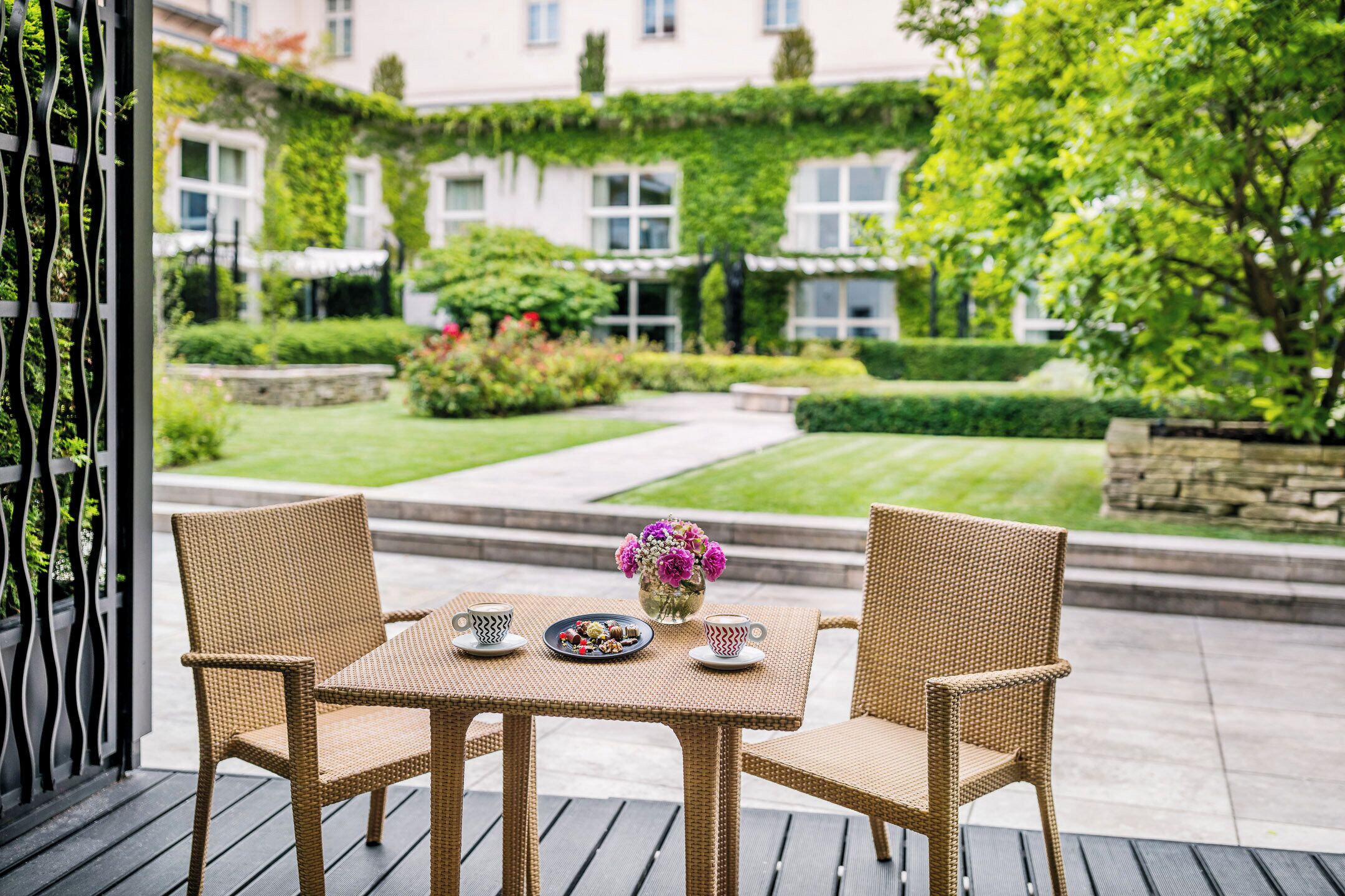 A wicker table set with chairs, a plate of pastries, and a vase of flowers on a balcony overlooking a well-manicured garden.