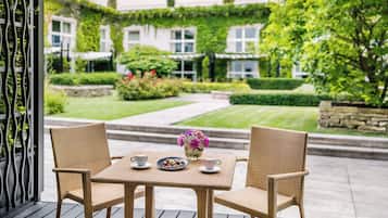 A wicker table set with chairs, a plate of pastries, and a vase of flowers on a balcony overlooking a well-manicured garden.