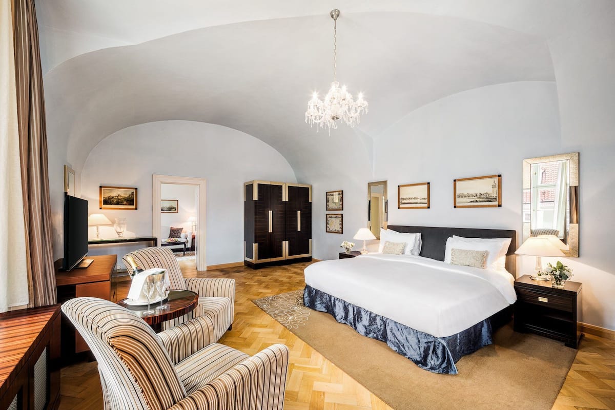 A spacious hotel room with a large bed, a sitting area with striped chairs, a television, and a chandelier.
