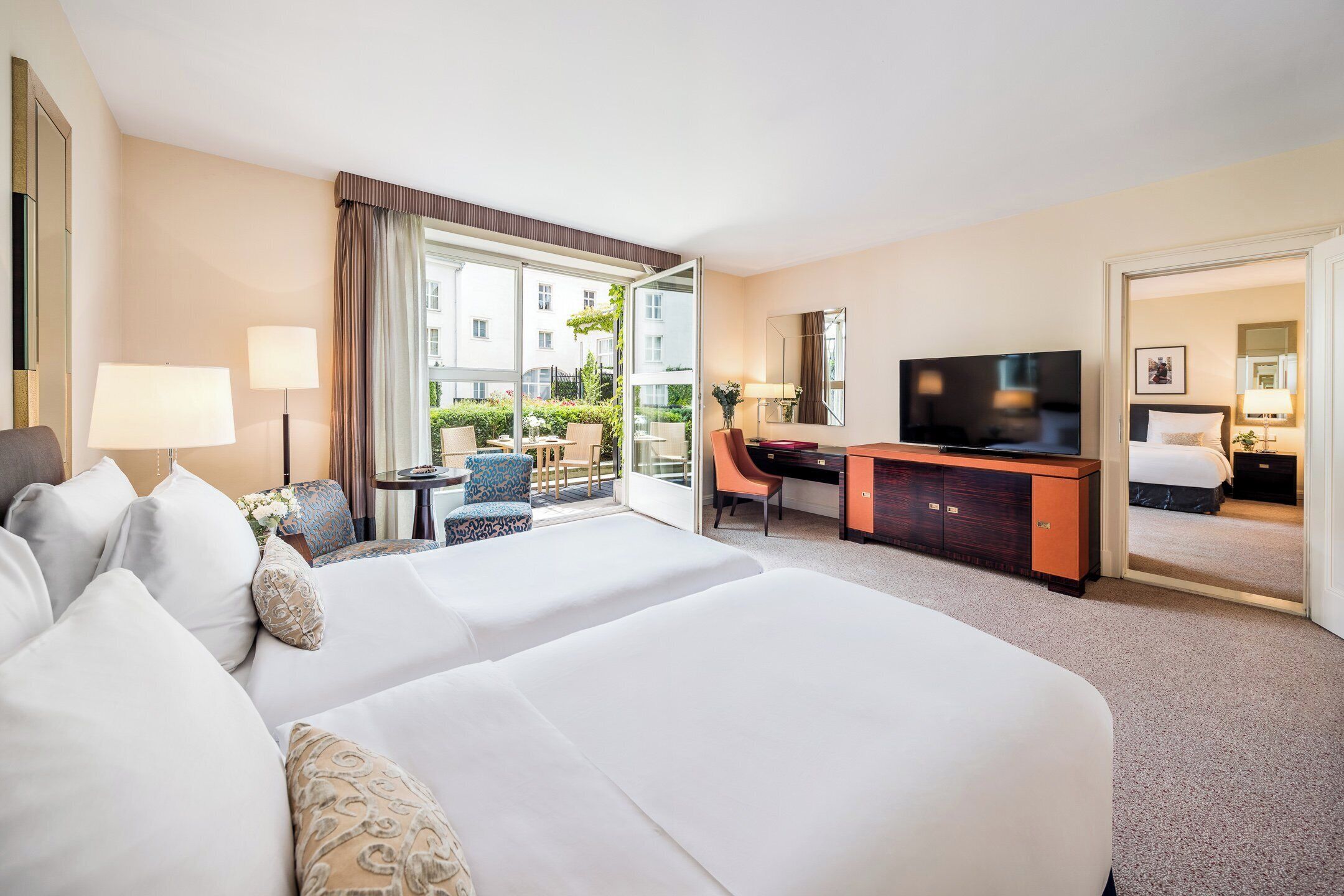 A hotel room with two beds, a desk, a television, and a view of the outside.