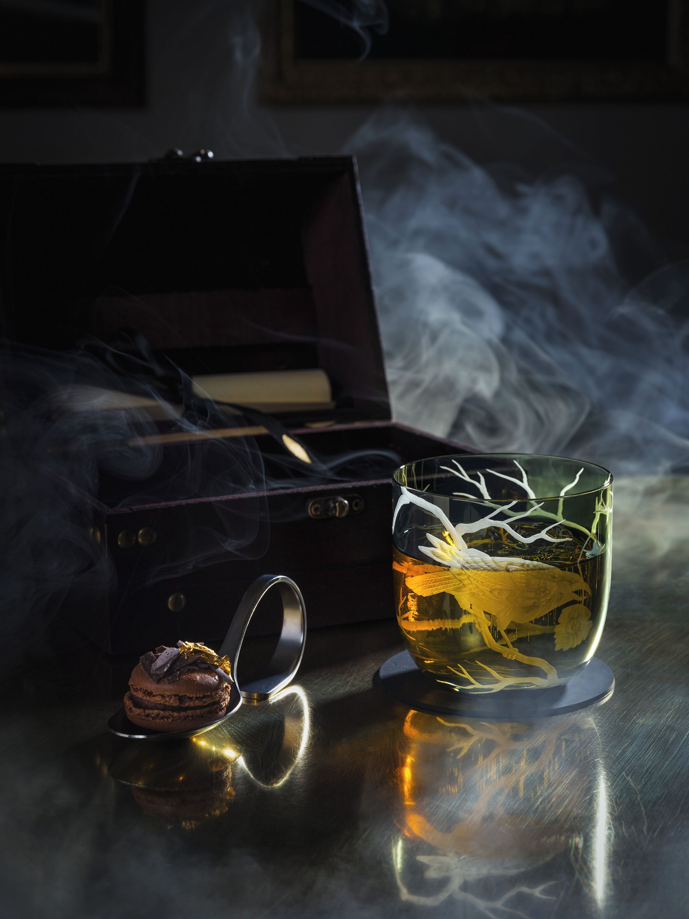 A dark room with a smoking pipe, a glass of tea with a flower, and a chocolate dessert on a reflective surface.