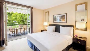A hotel room with a large bed, two bedside tables, a nightstand, a chair, a table, and a view of a garden through an open door.