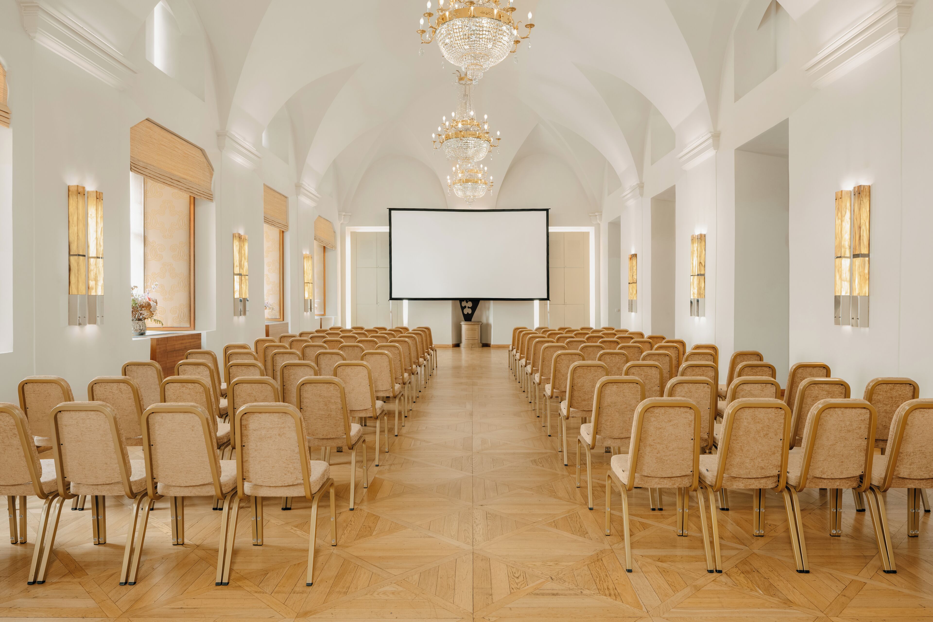 Ett konferensrum med rader av stolar, en stor skärm och kristallkronor.