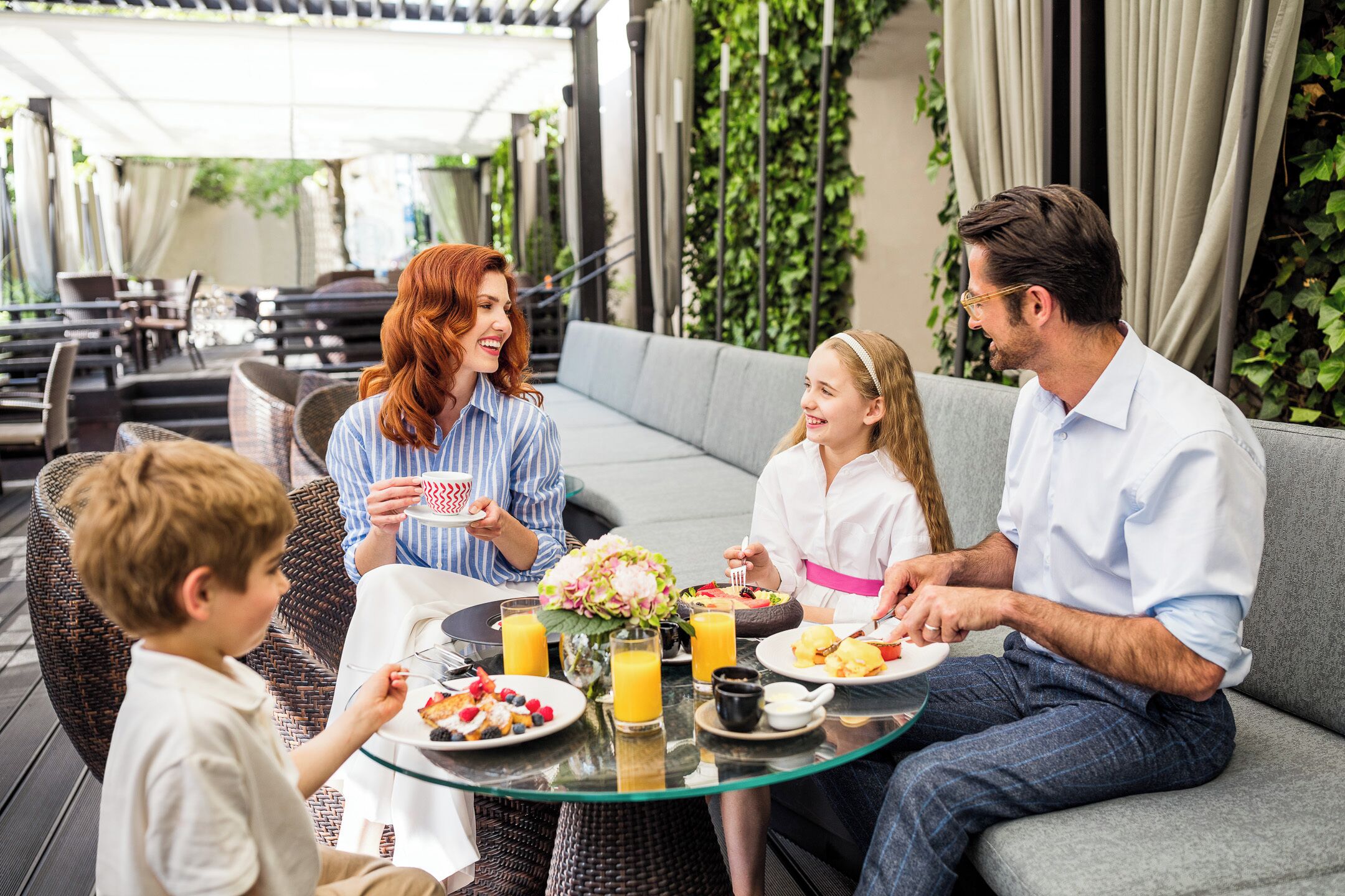 A family dining outdoors with a glass table, wicker chairs, and a grey sofa.