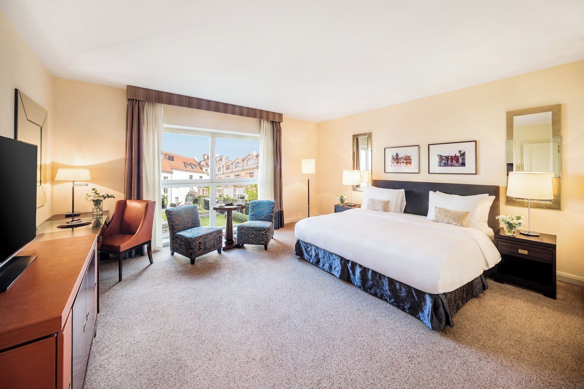 A hotel room with a large bed, a desk with a TV, two armchairs, a small table, and a view of buildings outside.