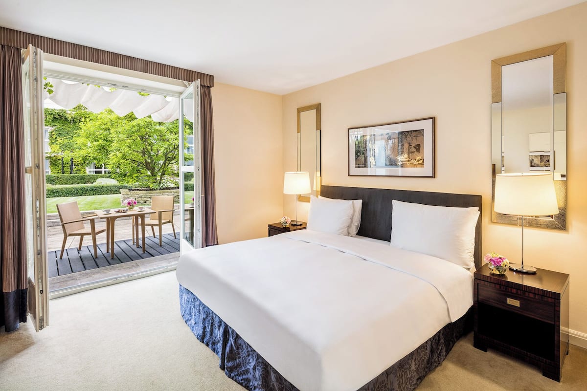 A hotel room with a large bed, two bedside tables, a nightstand, a chair, a table, and a view of a garden through an open door.