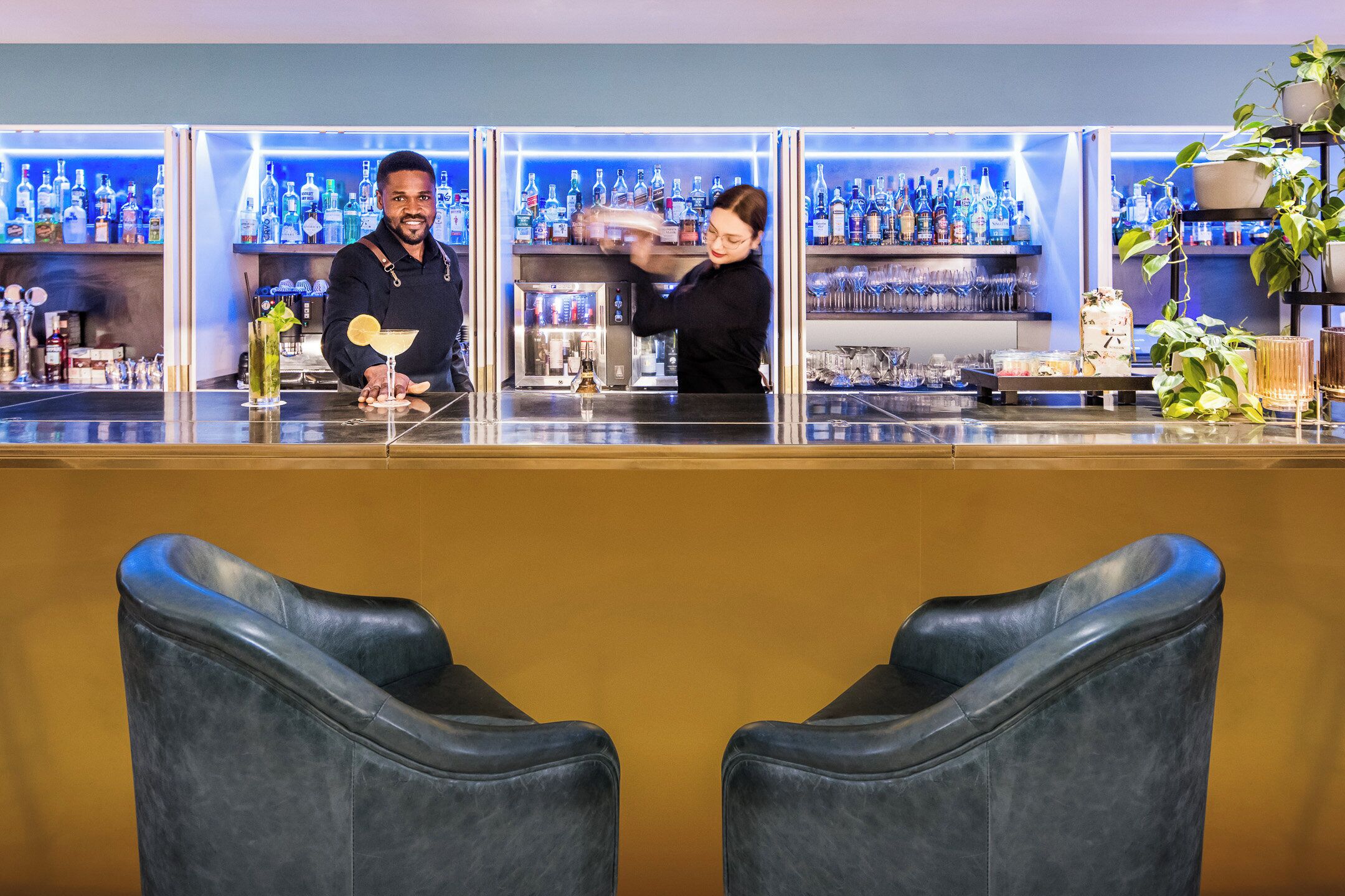 Eine Bar mit zwei Barhocker, einem Barkeeper und einer gut sortierten Flaschenauswahl.