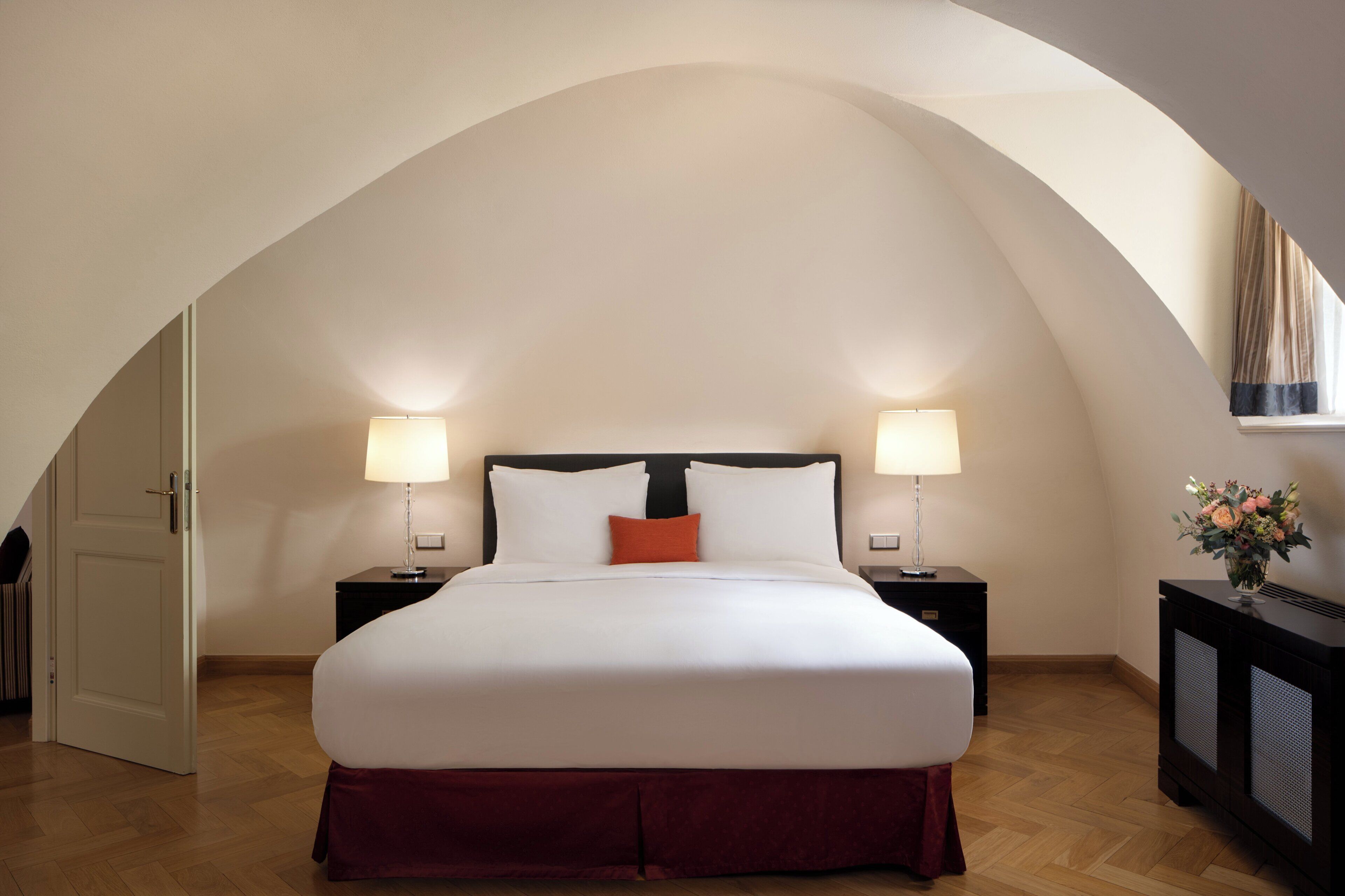 A bedroom with a large bed, two bedside tables with lamps, a nightstand, and a window with curtains.
