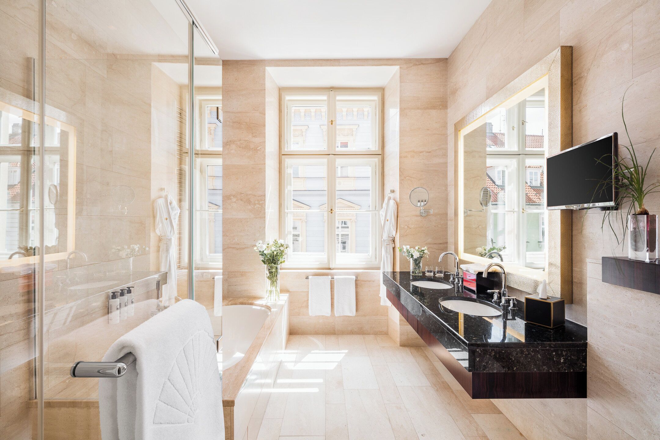 A modern bathroom with a large window, a bathtub, a double sink vanity, and a television mounted on the wall.