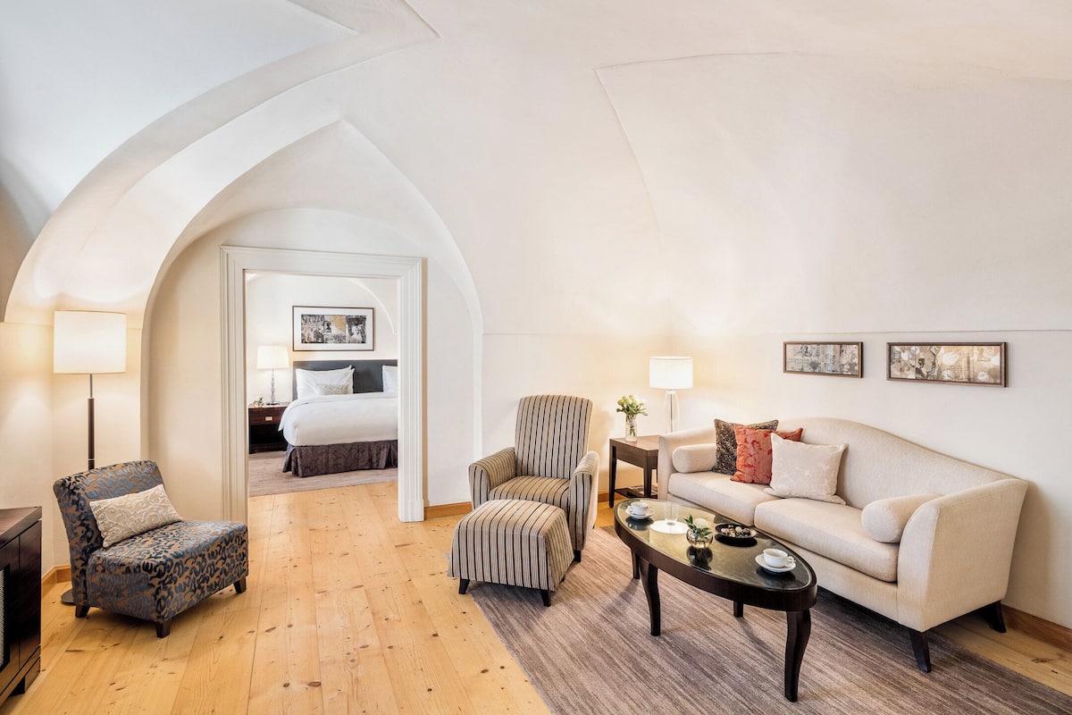 A hotel room with a wooden floor, a sofa, a chair, a coffee table, and a bed with a nightstand.