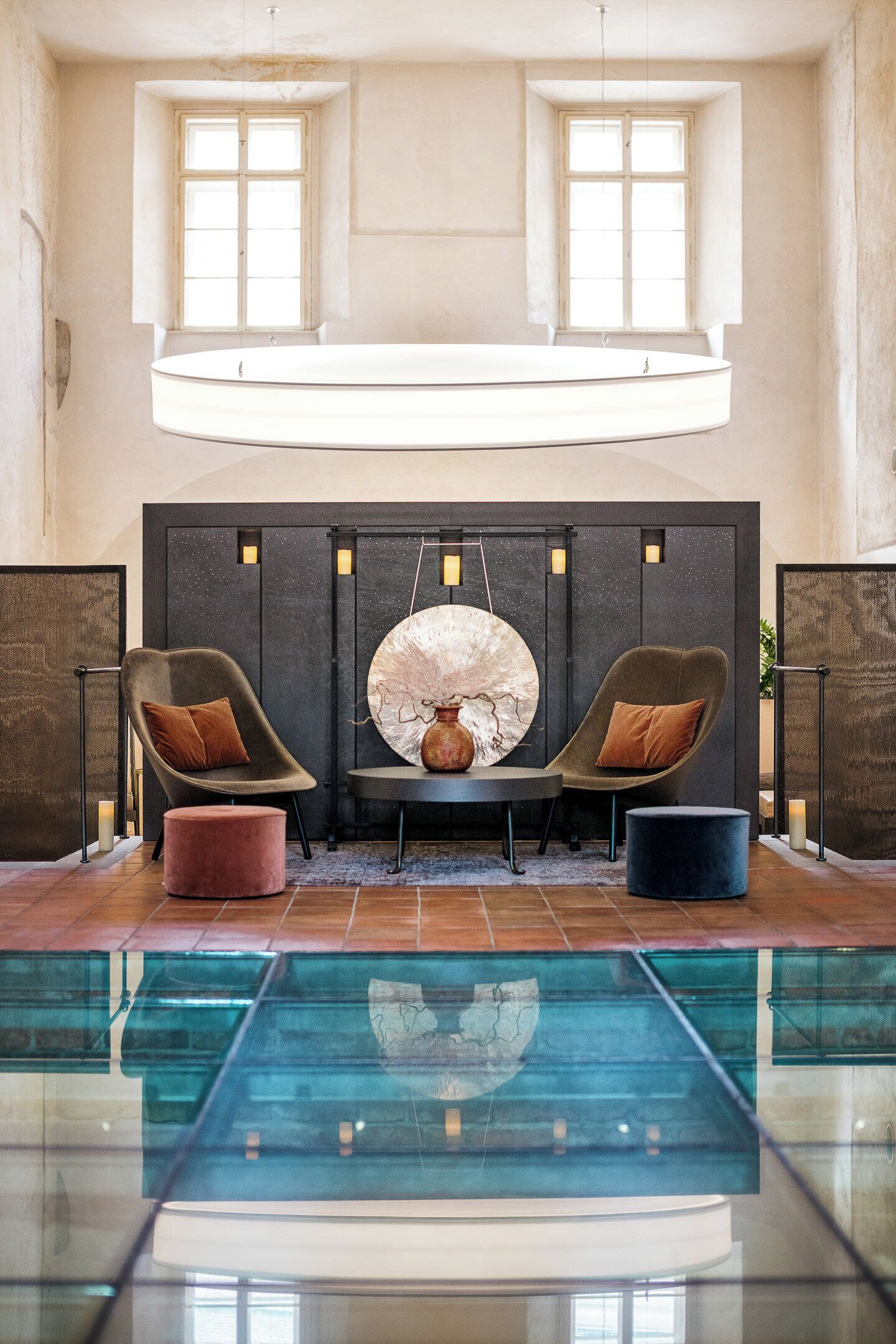 A pool area with two lounge chairs, a round table with a vase, and a wall with a circular decorative piece.