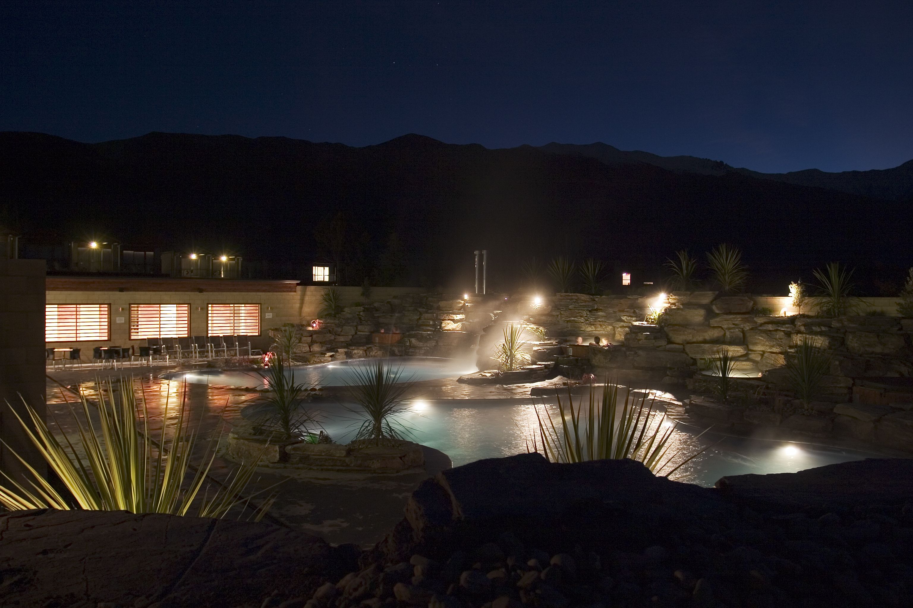 A nighttime view of a hotel with a pool area, stone walls, and a mountain backdrop.