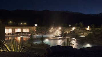 A nighttime view of a hotel with a pool area, stone walls, and a mountain backdrop.