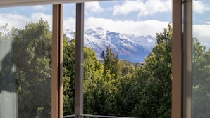 Vista pela janela mostrando uma paisagem montanhosa, com picos nevados e árvores verdes densas.