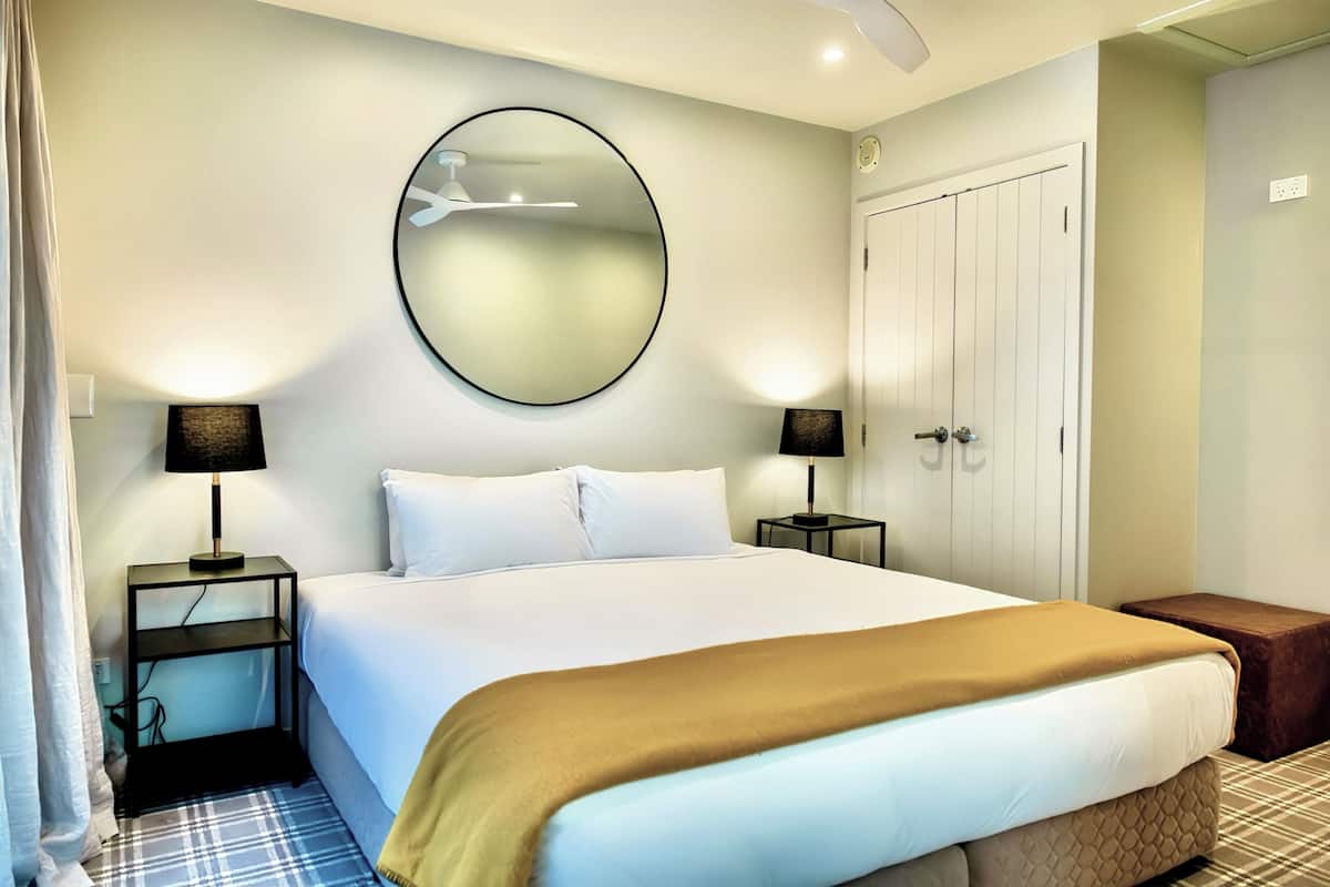A hotel room with a large bed, two bedside tables with lamps, a round mirror on the wall, a wardrobe, and a small bench.