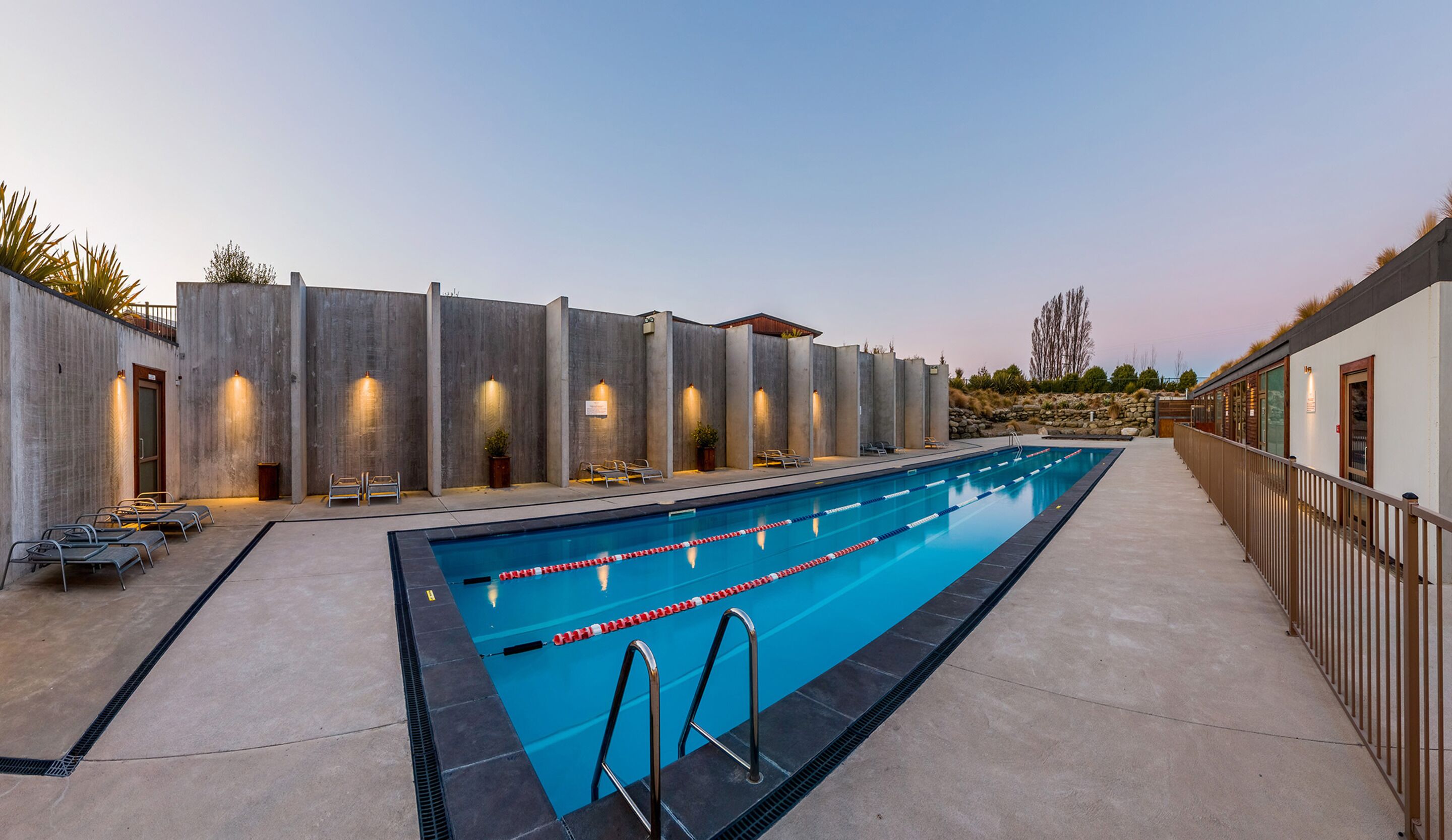 En swimmingpool med en betongplatta runt om, solstolar och en stenmur med inbyggda lampor.