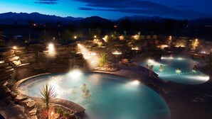 Vista noturna de um resort com várias banheiras de hidromassagem e piscinas, cercado por muros de pedra e iluminado por iluminação ambiente.