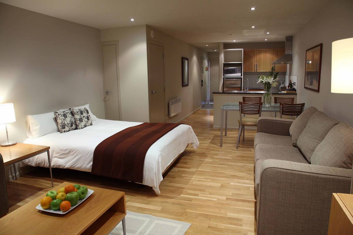 A hotel room with a bed, a sitting area with a sofa, a dining table with chairs, and a kitchen area in the background.