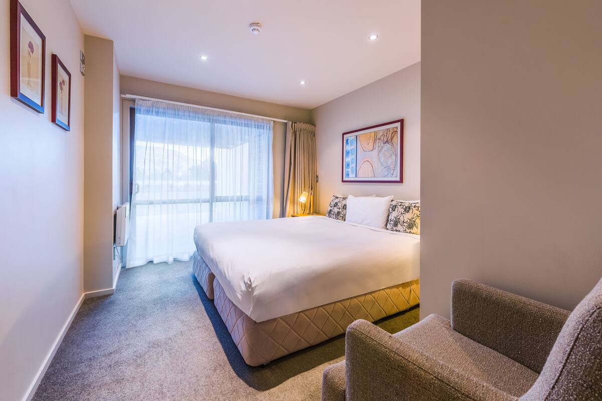 A hotel room with a large bed, a chair, a painting on the wall, and a window with curtains.