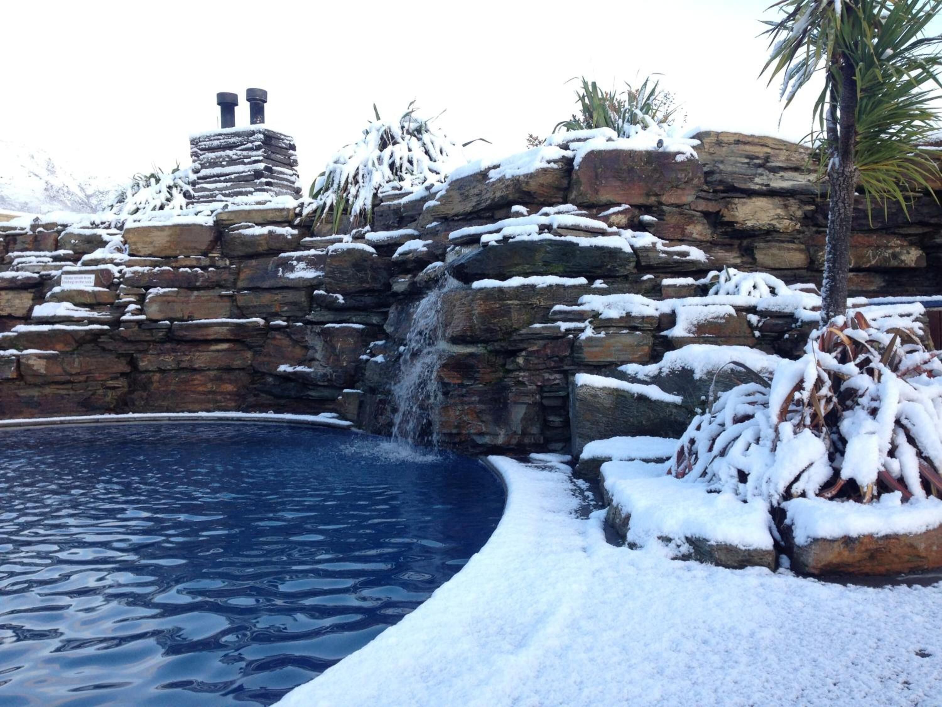 A pool with a waterfall feature surrounded by snow-covered rocks and a palm tree.
