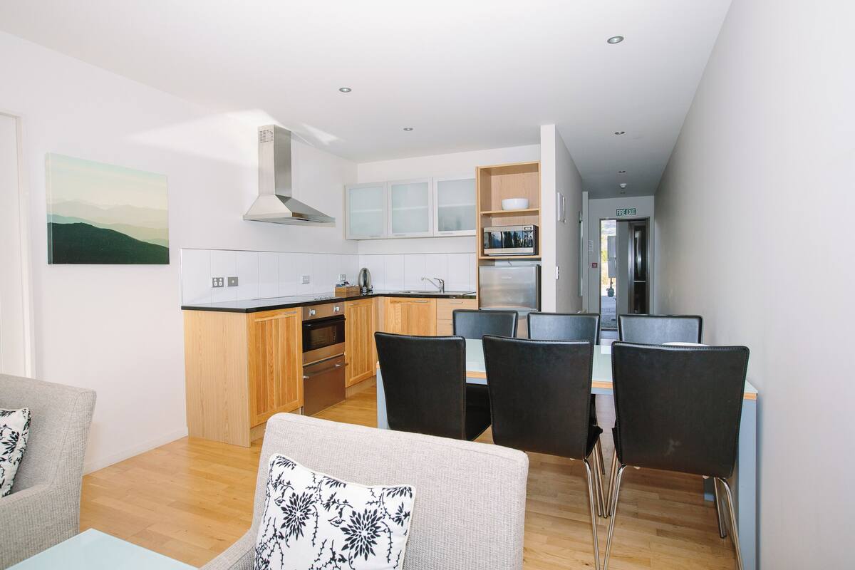 A modern kitchen with wooden cabinets, a built-in microwave, and a stainless steel range hood. There is a dining area with a table and chairs, and a sofa with a patterned cushion.