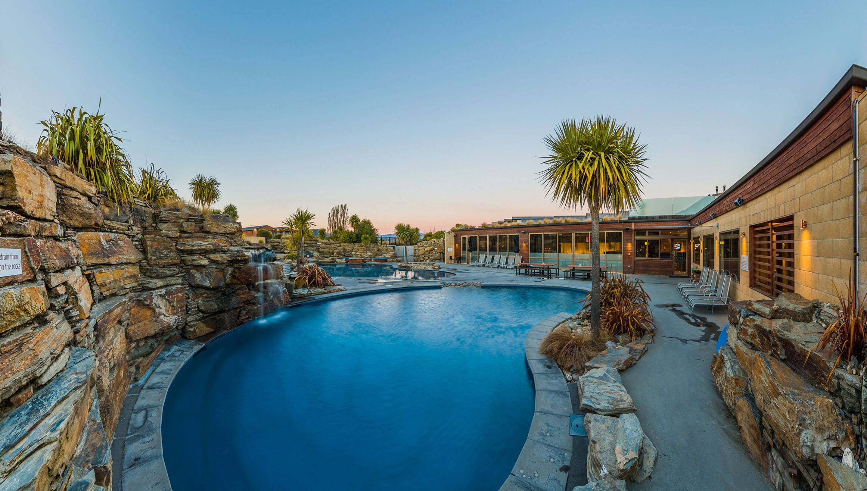 A swimming pool with a stone wall, palm trees, and a building with outdoor seating.