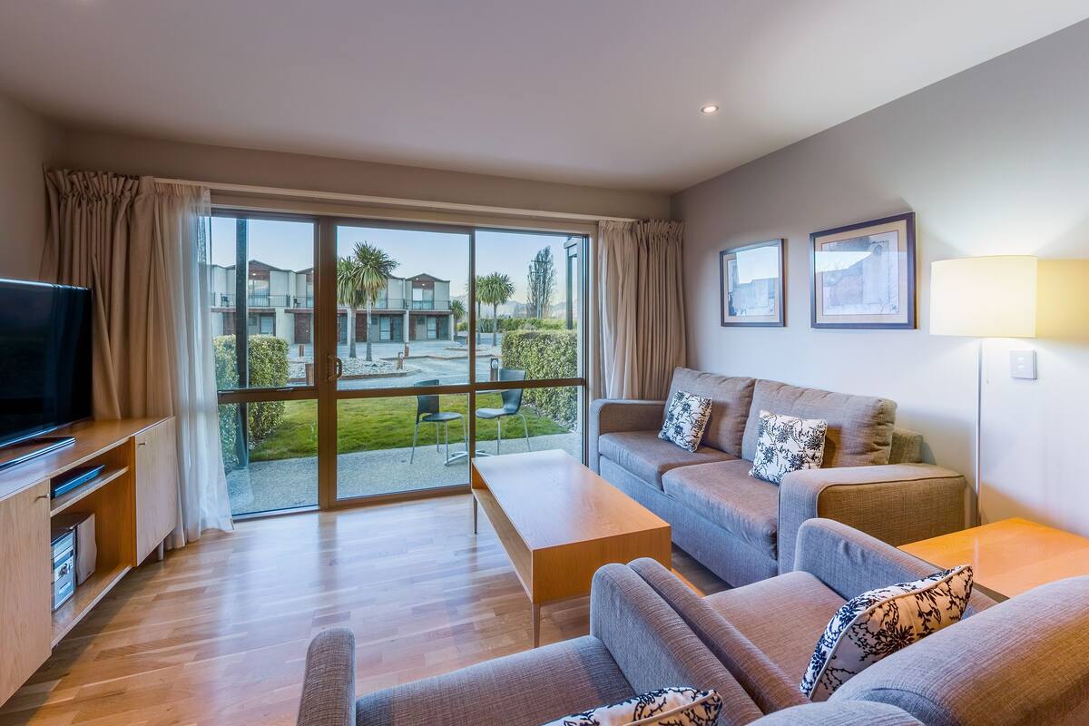 A modern living room with a sofa set, a coffee table, and a flat-screen TV. Large windows offer a view of a courtyard with palm trees and a building.
