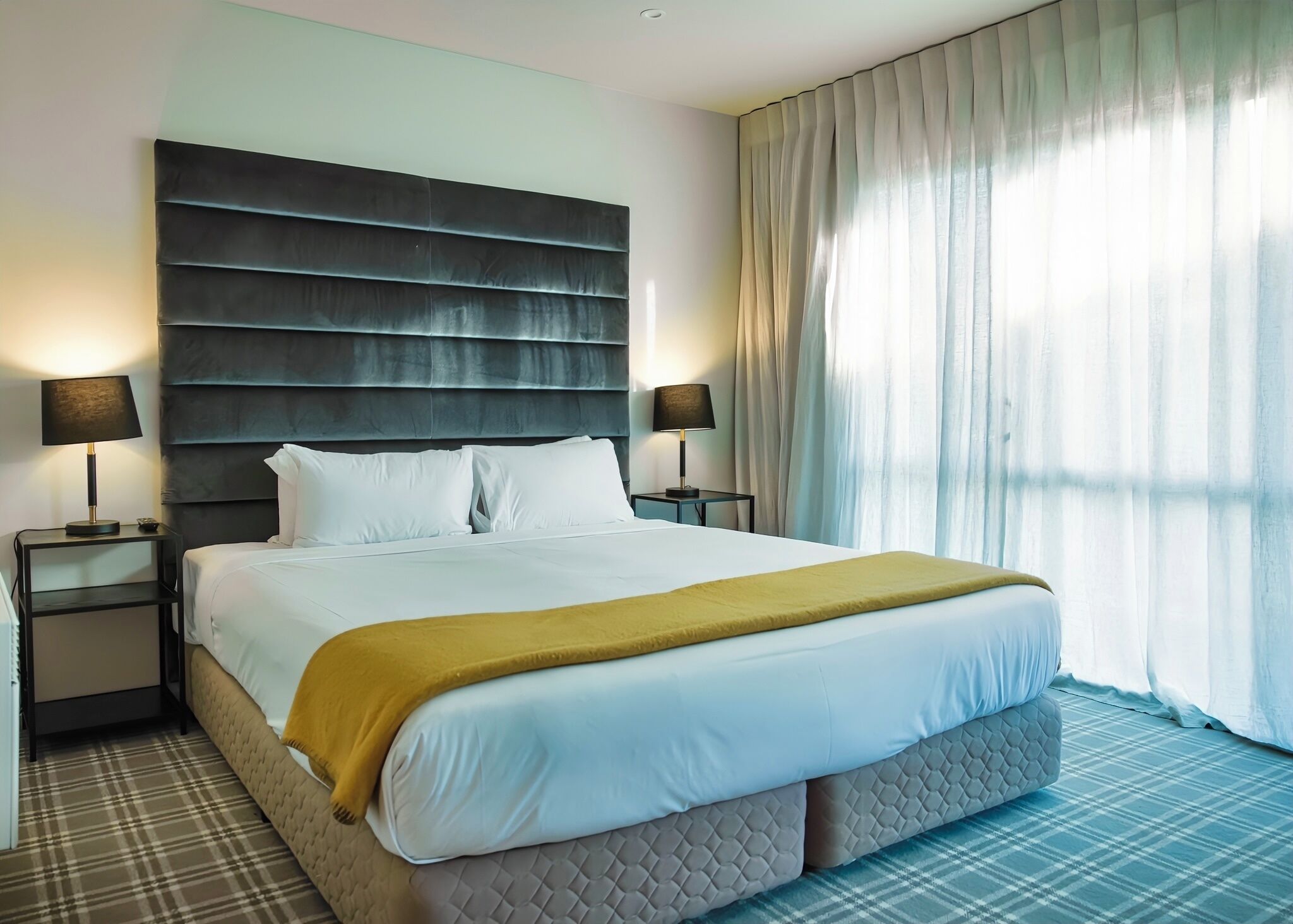 A hotel room with a large bed, two bedside tables with lamps, a headboard, and a window with curtains.