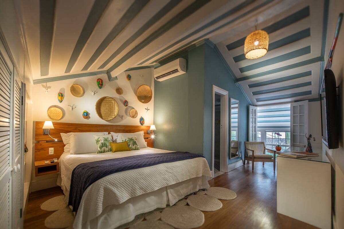 A bedroom with a bed, bedside tables, a desk, and a chair. There is a window and a door leading to another room.