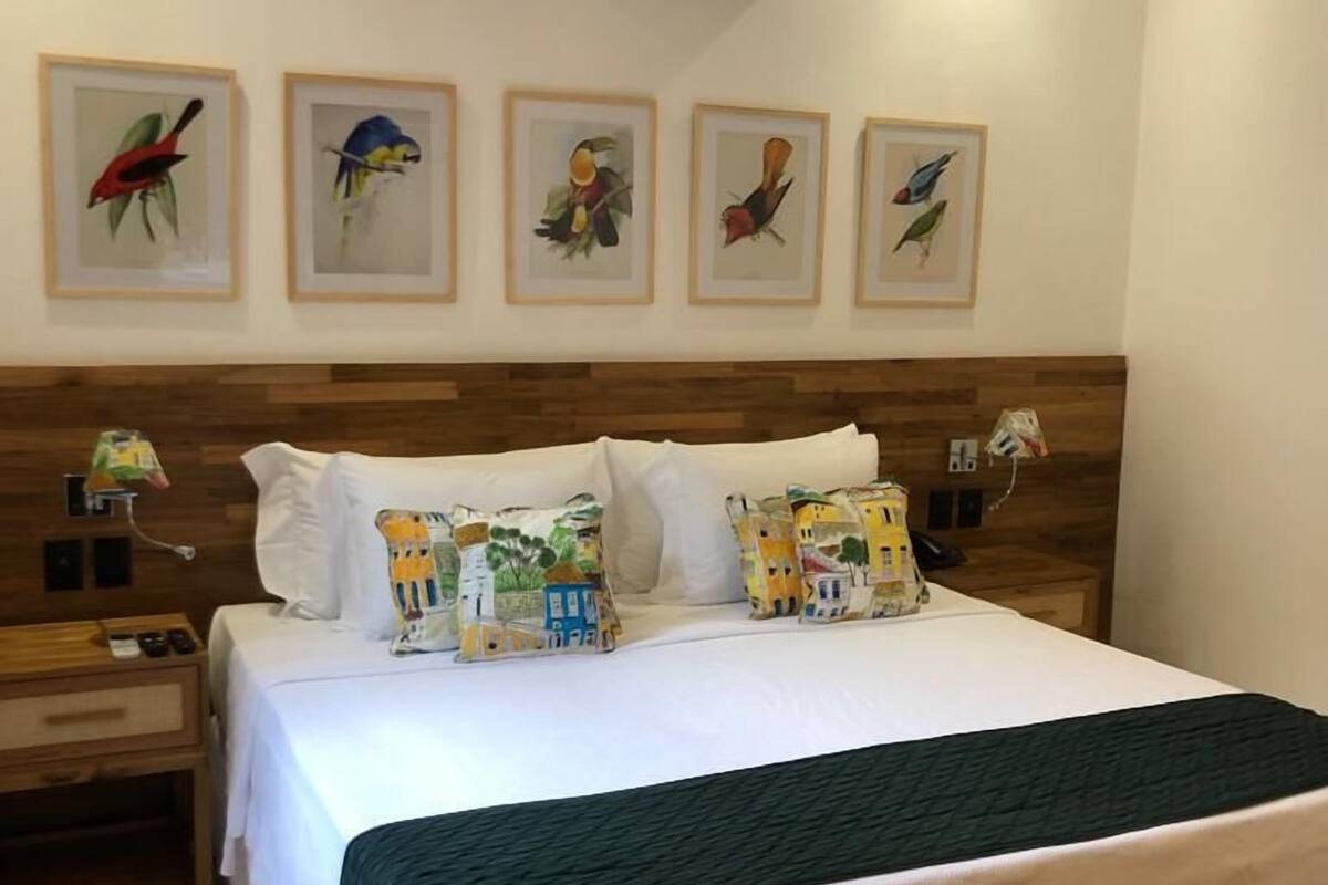 A hotel room with a large bed, wooden headboard, multiple pillows, a bedside table, a nightstand, a bench, and wall-mounted air conditioning.