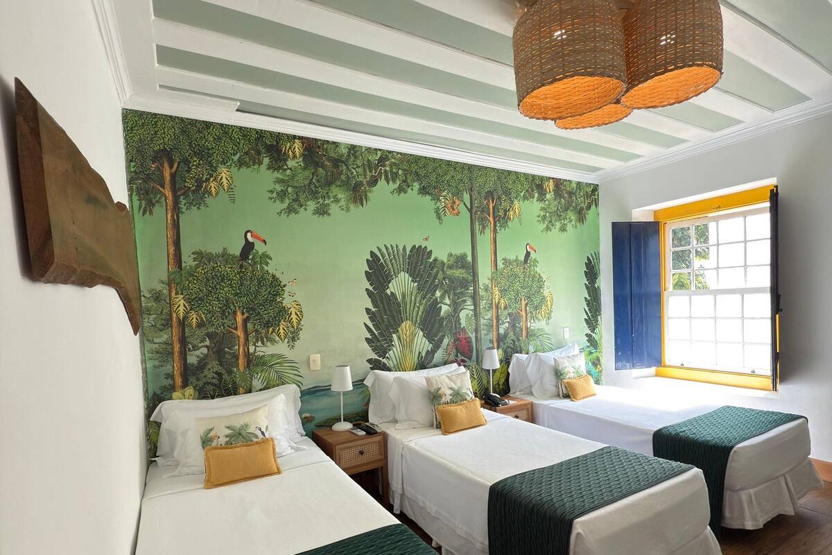 A room with three beds, a mural of a forest, a window with yellow blinds, and a woven light fixture.