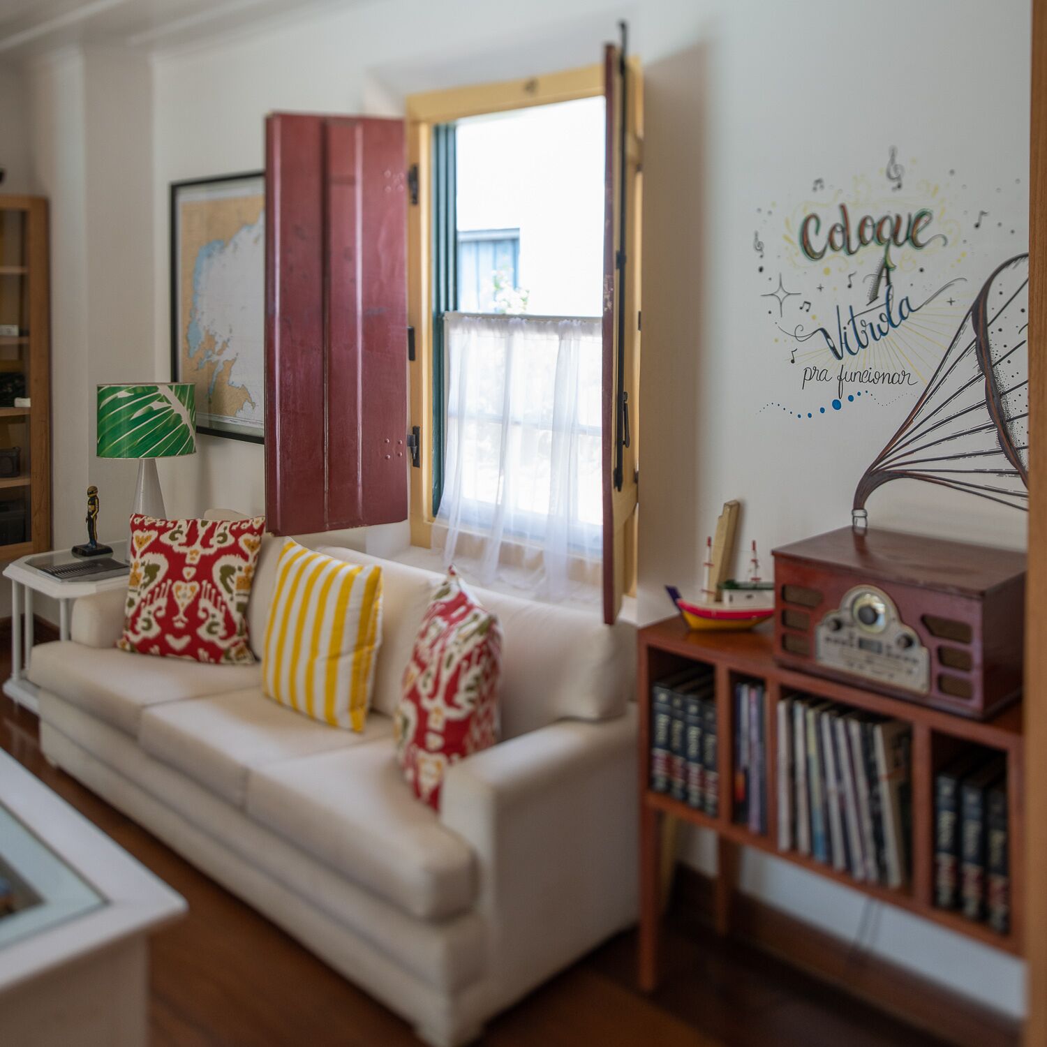 Una sala de estar con un sofá blanco, cojines estampados, una mesita con una lámpara, una estantería y una pared con un letrero decorativo.