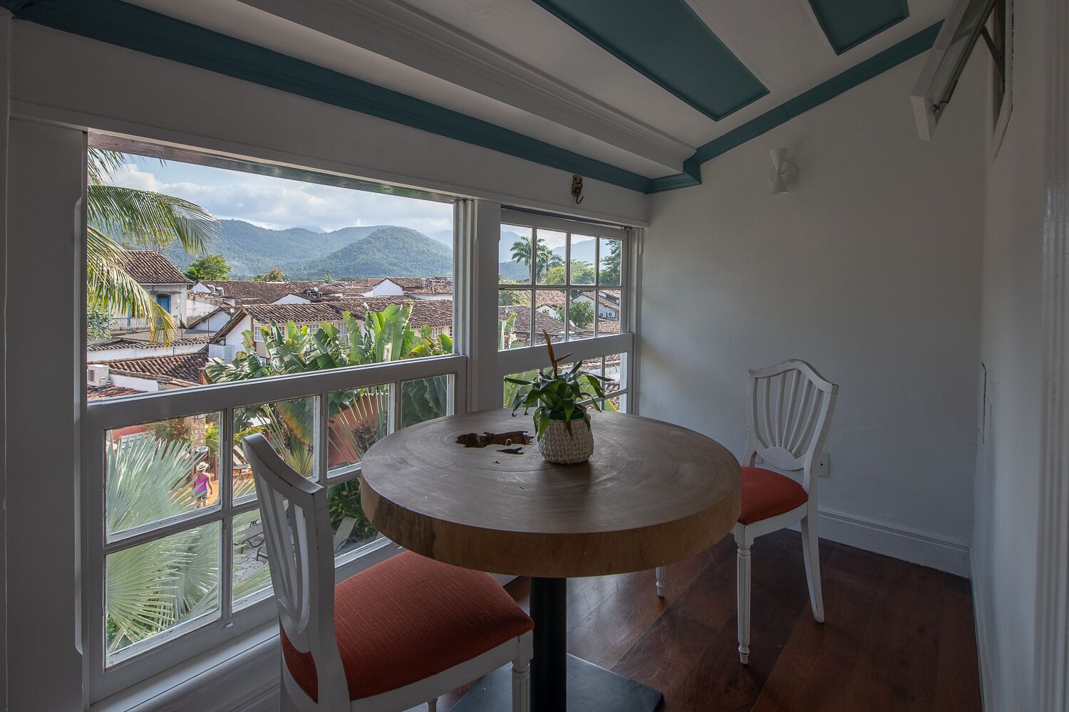 Quarto com uma mesa de madeira redonda, duas cadeiras brancas e vista para uma paisagem montanhosa através de grandes janelas.