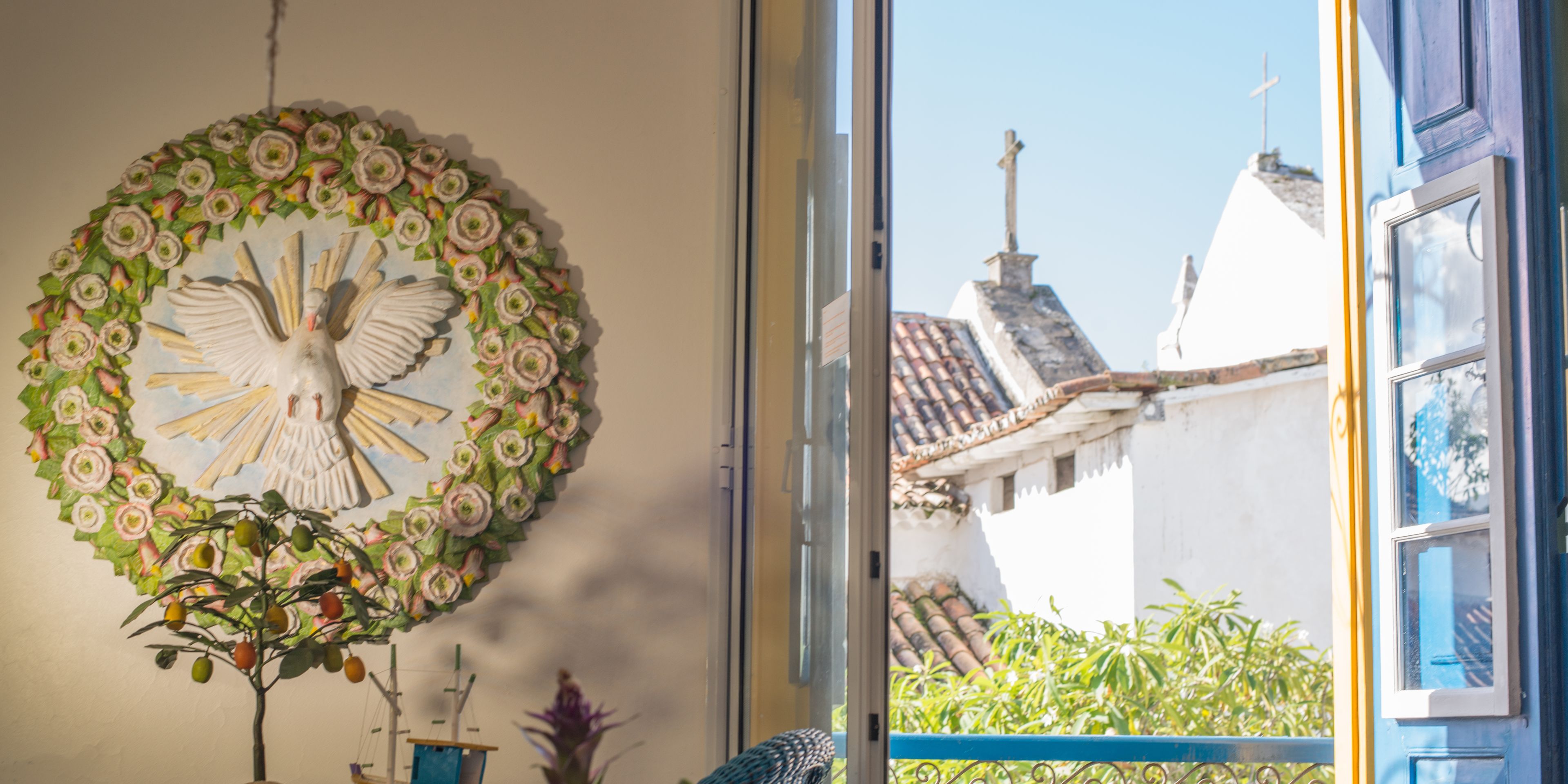 A room with a large window, a decorative wreath on the wall, and a view of buildings with a cross on top.