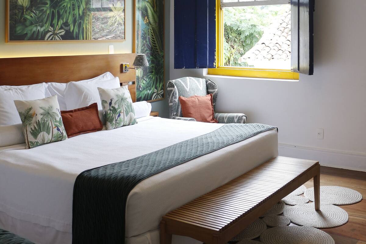 A bedroom with a large bed, a bench, a window with yellow shutters, and tropical-themed artwork.