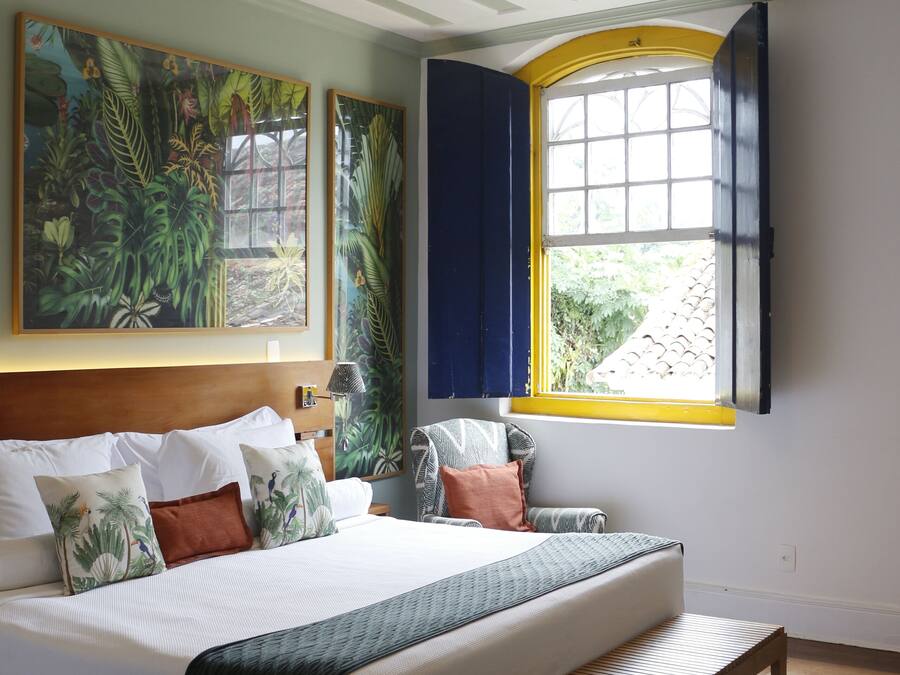 Un dormitorio con una cama grande, un banco, una ventana con contraventanas amarillas y obras de arte con temática tropical.