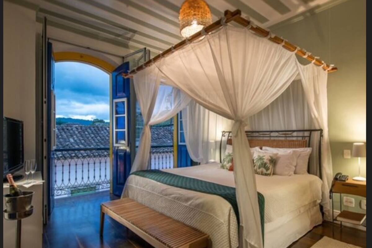 A bedroom with a canopy bed, a bench, a bedside table, and a view of the outside balcony.