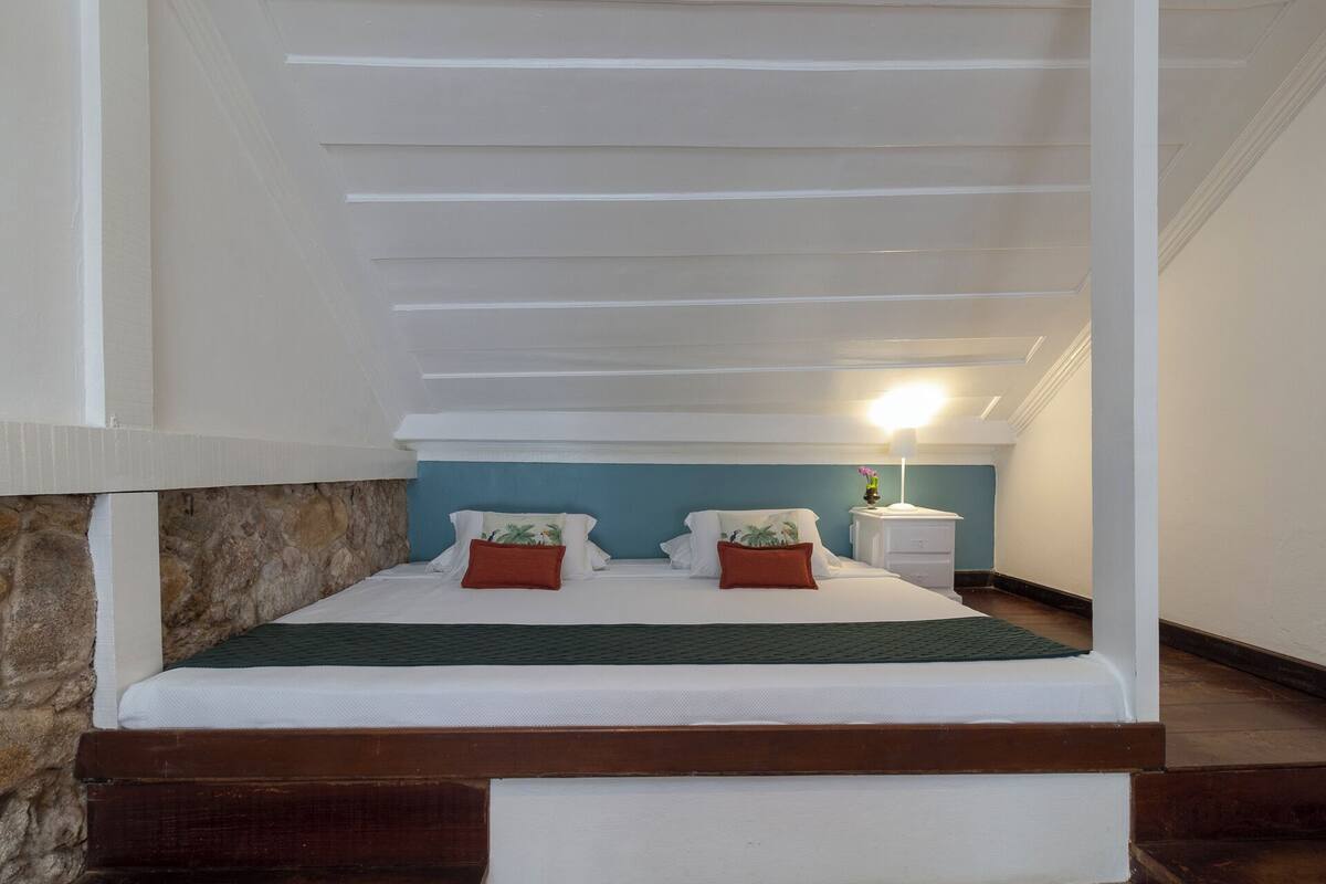 A bedroom with a sloped ceiling, a stone wall, a bed with white bedding and two pillows, a nightstand with a lamp, and a blue accent wall.